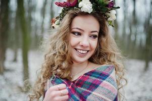 Curly cute blonde girl with wreath in checkered plaid at snowy forest in winter day. photo