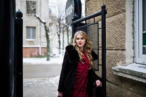 Elegance blonde girl in red evening dress and fur coat at streets of city in winter day. photo