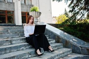 Good-looking young woman in white blouse, wide black pants and black classic high heels sitting on stairs and working on her laptop. photo