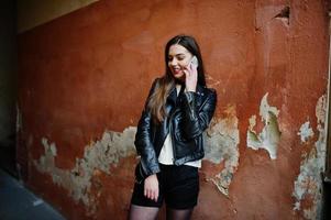 Stylish brunette girl wear on leather jacket and shorts with mobile phone at hands against orange wall. photo