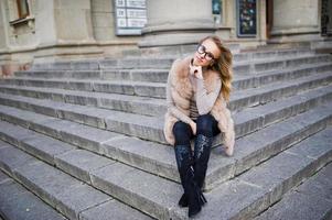 chica rubia en abrigo de piel y gafas sentada en las escaleras. foto