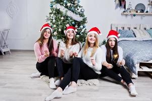 Four cute friends girls wear on warm sweaters,  black pants and red santa hats against new year tree with christmas decoration at white room and hold bengal lights at hands. photo