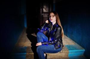 Night portrait of girl model wear on glasses, jeans and leather jacket, with blue garland on her. photo