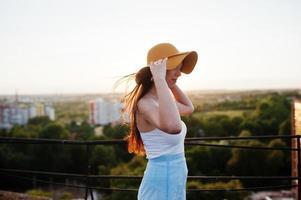 Portrait of a pretty young woman in white t-shirt and blue skirt posing on the rooftop with her orange hat at the sunset. photo