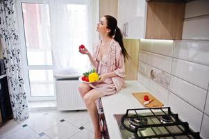 retrato de una bella joven vestida de rosa posando con un tomate en su cocina. foto