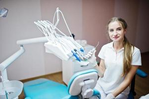 Una dentista guapa posando con algunos instrumentos dentales en la mano con bata blanca en un gabinete moderno y bien equipado. foto