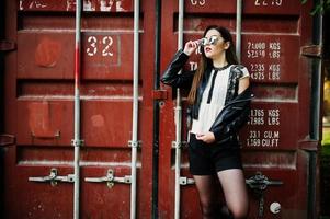 Stylish brunette girl wear on leather jacket and shorts, sunglasses against shipping container. photo