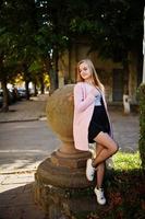 Young blonde girl in black skirt and pink coat posed at sunny day on street. photo