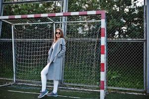 Girl in gray coat with sunglasses at small street stadium football gates. photo