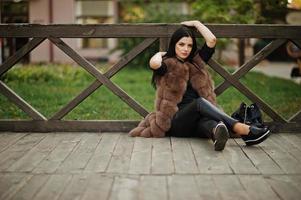 Fashion outdoor photo of gorgeous sensual woman with dark hair in elegant clothes and luxurious fur coat sitting against wooden railings.