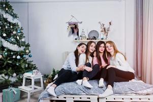 Four cute friends girls wear on warm sweaters and black pants on bed at new year decorated room on studio. photo