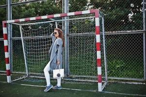 Girl in gray coat with sunglasses at small street stadium football gates. photo