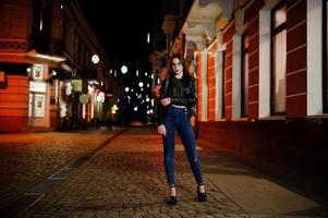 retrato nocturno de modelo de niña con gafas, jeans y chaqueta de cuero, con mochila, contra las luces de las calles de la ciudad. foto