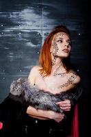Fashion model red haired girl with originally make up like leopard predator against steel wall. Studio portrait on ladder. photo