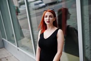 Red haired stylish girl in sunglasses wear in black, against large windows. Street fashion portrait. photo