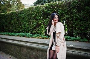 Black hair sexy woman in glasses and coat posed on street with mobile phone.