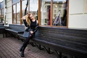 Blonde fashionable girl in long black leather coat sitting on bench. photo