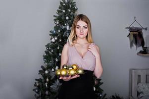 Blonde girl in dress posed near new year tree with christmas gifts decor in white room. photo