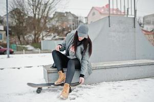 chica morena elegante con gorra gris, estilo casual de calle con patineta en el día de invierno. foto