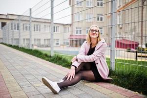 Blonde girl at glasses and pink coat, black tunic sitting against fence at street. photo