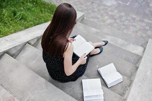 Portrait of a fabulous woman in black polka dot dress sitting on the stairs and reading in the park. photo