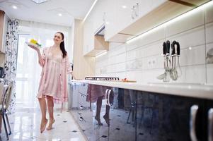 chica atractiva en túnica rosa posando con un plato de ensalada en la cocina. foto