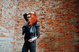 chica elegante de pelo rojo con gafas de sol vestidas de negro, contra un lugar abandonado con paredes de ladrillo. foto