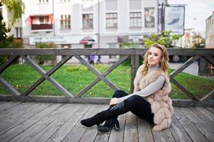 Blonde girl at fur coat sitting on wooden floor outdoor. photo