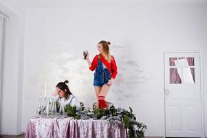 Two fun beautiful girls friends wear in overalls jeans shorts and gaiters against table with new year decoration. photo