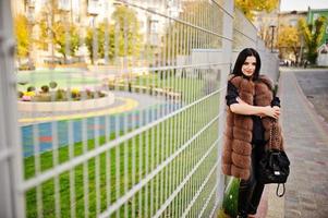 Fashion outdoor photo of gorgeous sensual woman with dark hair in elegant clothes and luxurious fur coat and with backpack against iron fence.
