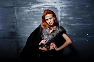 Fashion model red haired girl with originally make up like leopard predator wear on furs against steel wall. Studio portrait.