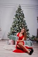 Fashionable gorgeous woman at red long evening dress posed at studio against new year tree with gifts. Christmas holidays theme. photo
