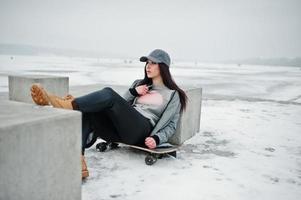 Stylish brunette girl in gray cap, casual street style with skate board on winter day. photo