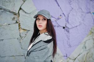 chica morena elegante con gorra gris, estilo casual de calle en el día de invierno contra la pared de color. foto