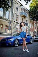 Curly stylish girl wear on blue jeans skirt, blouse and glasses posed near blue car at street of city. photo