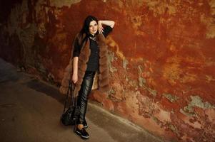 Fashion outdoor photo of gorgeous sensual woman with dark hair in elegant clothes and luxurious fur coat at old street with grunge walls.