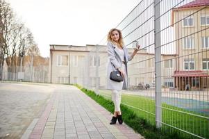 Stylish curly blonde model girl wear on white posing against fence. photo