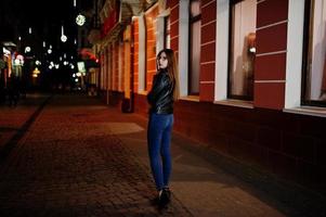 retrato nocturno de modelo de niña con gafas, jeans y chaqueta de cuero, con mochila, contra las calles de la ciudad. foto