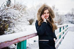 Beautiful brunette girl in winter warm clothing. Model on winter jacket against frozen lake at park speak on mobile phone. photo