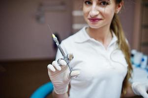 Portrait of a female dentist in white coat posing with syringe filled with anesthesia in a dental cabinet. photo
