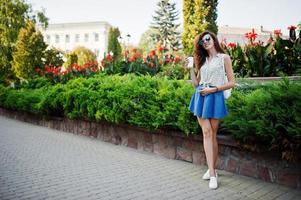 ropa de niña con estilo rizado en falda de jeans azules, blusa y gafas posadas en la calle de la ciudad con una taza de café. foto