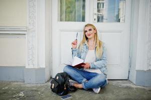 una chica rubia usa jeans con mochila posada contra una puerta vieja con un diario y escribe algo. foto