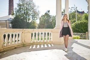 Young blonde girl in black skirt and pink coat posed against vintage railing with a column of old house. photo