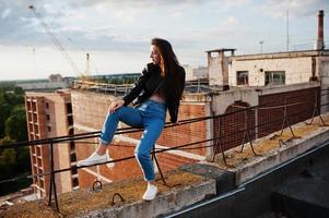 retrato de una hermosa joven con chaqueta de cuero negro, jeans y zapatillas sentadas en pasamanos en el techo con vistas pintorescas a un parque. foto