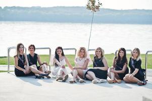 Group of 7 girls wear on black and 2 brides sitting background lake at hen party. photo