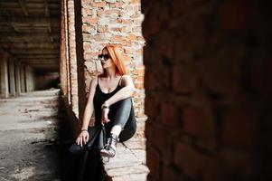 chica elegante de pelo rojo con gafas de sol vestidas de negro, contra un lugar abandonado con paredes de ladrillo. foto