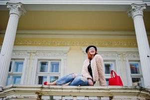 chica pelirroja con gafas y sombrero con bolso rojo posado cerca de la casa vintage. foto