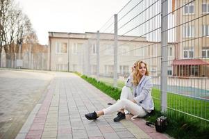 Stylish curly blonde model girl wear on white posing against fence. photo