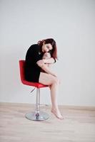 Red haired girl on black dress tunic sitting on red chair against white wall at empty room. photo