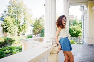 ropa de niña con estilo rizado en falda de jeans azules, blusa y gafas posadas cerca de la antigua casa vintage. foto